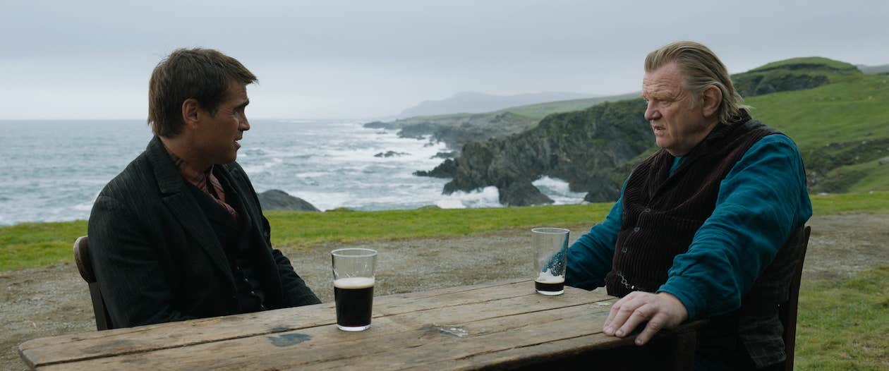 Brendan Gleeson and Collin Farrell from 'The Banshees of Inisherin' sit at a table with a scenic background, drinking pints of Guinness in a scene from the film.