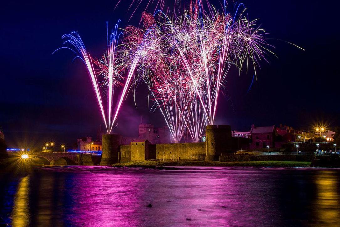 A night sky lit up by pink and white fireworks over an old castle on the bank of a river.