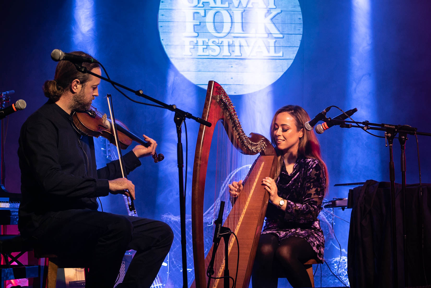 Brídín at Galway Folk Festival. A man playing the fiddle beside a woman playing the harp with mics on stands, with a lit up blue background with the festival logo in light blue.