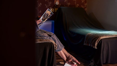 Bottom half of a person sitting on a chair holding a magazine with white slippers on the floor