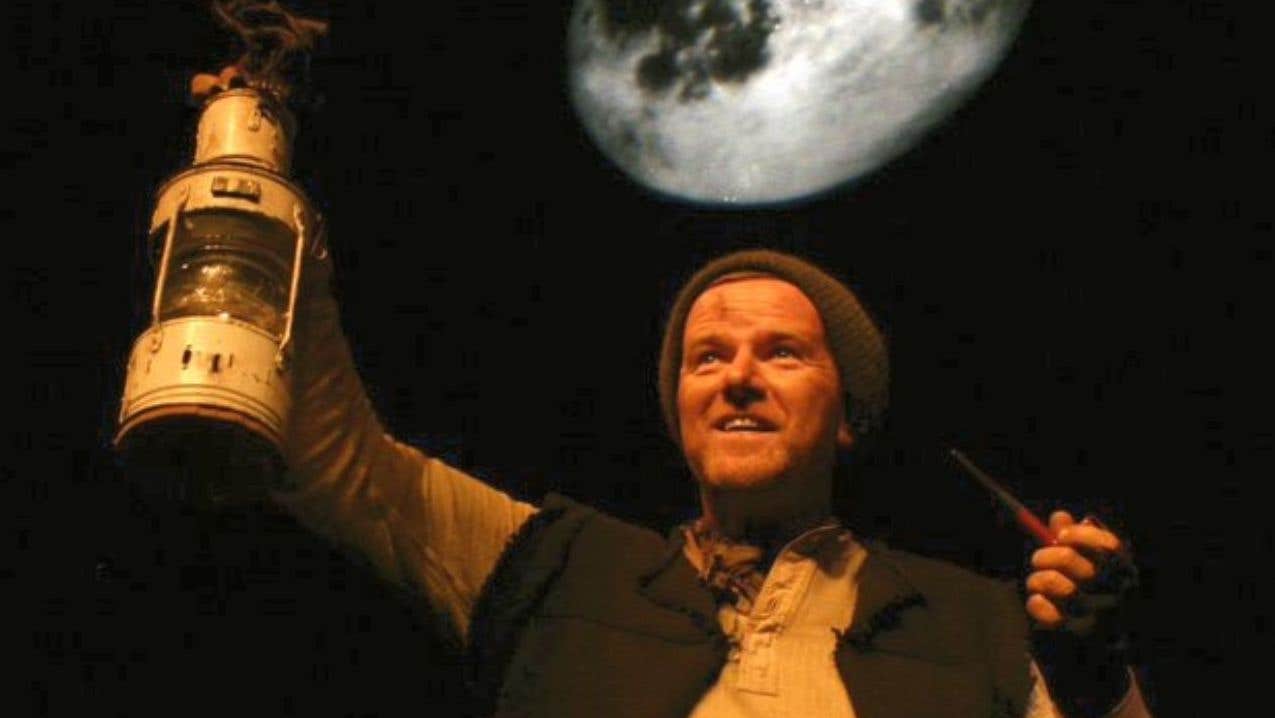 Against a dark background with a blurred, silvery moon, a man is holding up an old fashioned lamp, holding a pipe in his other hand, wearing a rough waistcoat and a woollen hat.