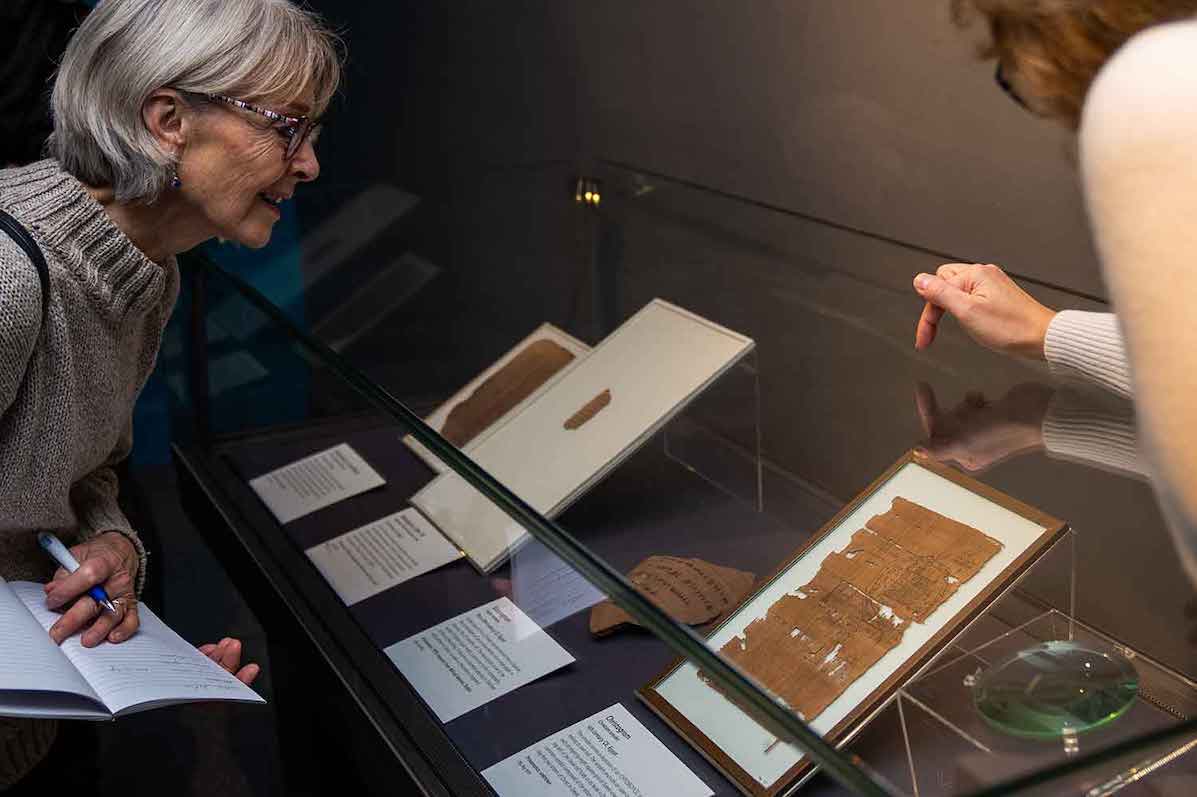 Photo of a woman on the left holding a notebook and pen looking at a glass display cabinet in centre of photo containing some old parts of manuscripts with display cards and the hand of another woman on the right pointing down.