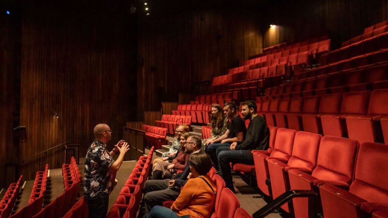 Small group of people sitting in a theatre auditorium with a tour guide