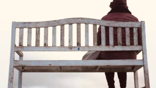 'An tÉalú' Ceoldráma Nua le Seán Óg Ó Duinnín - rear view of person in winter coat and hat seated at one end of a wooden bench.