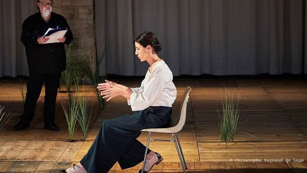 An image from Sopro. Two people on stage, one is sitting the foreground another is standing in the background with a script in hand.