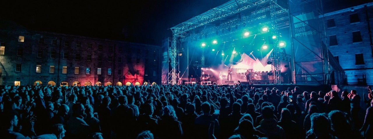 Distant view of large outdoor stage at night with large audience watching musicians.