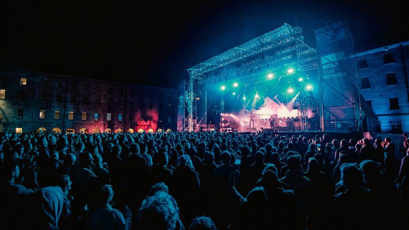 Distant view of large outdoor stage at night with large audience watching musicians.
