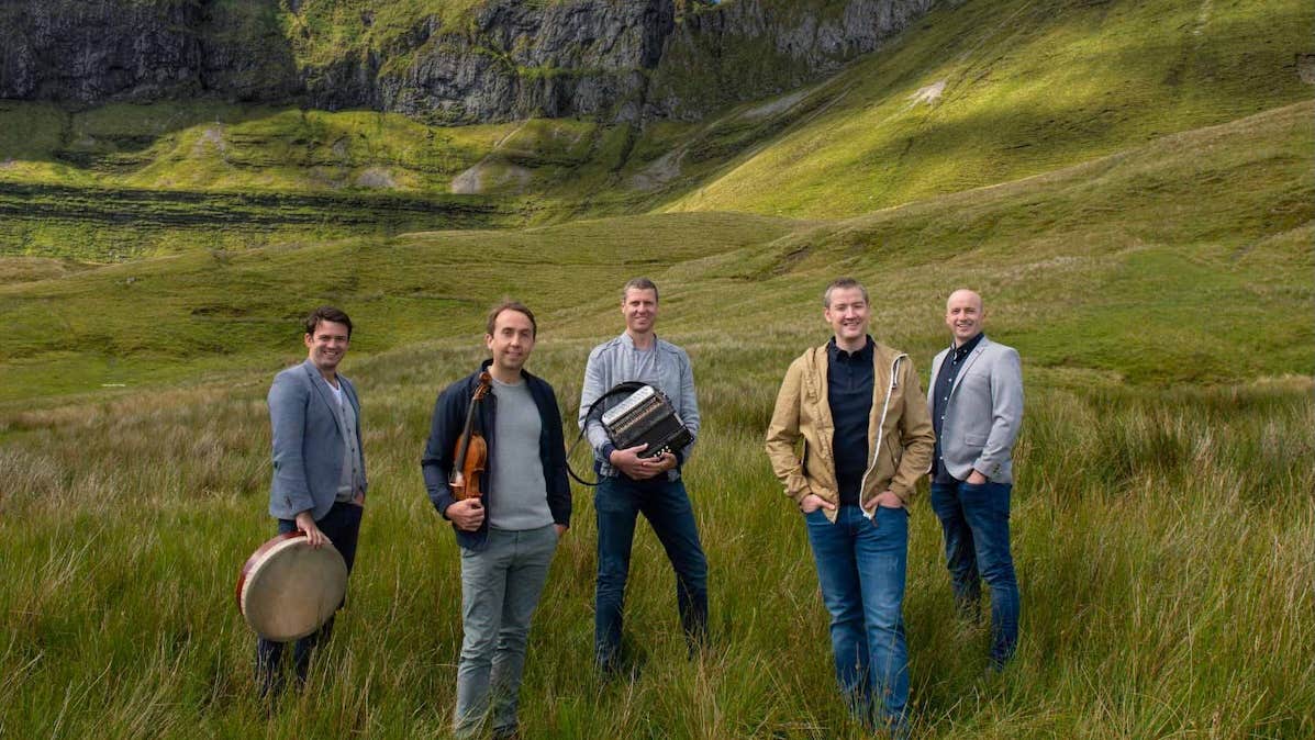 Group of 5 men standing in grassy area with backdrop of rocky hills