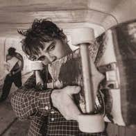 A close up of man holding up a skateboard horizontally to his shoulder.