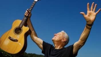 A man with arms outstretched, holding up a guitar in his right hand against blue sky