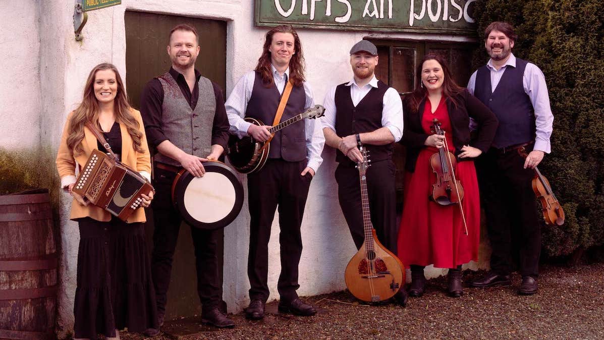 A line of 6 smiling men and women holding musical instruments standing in front of an old cottage