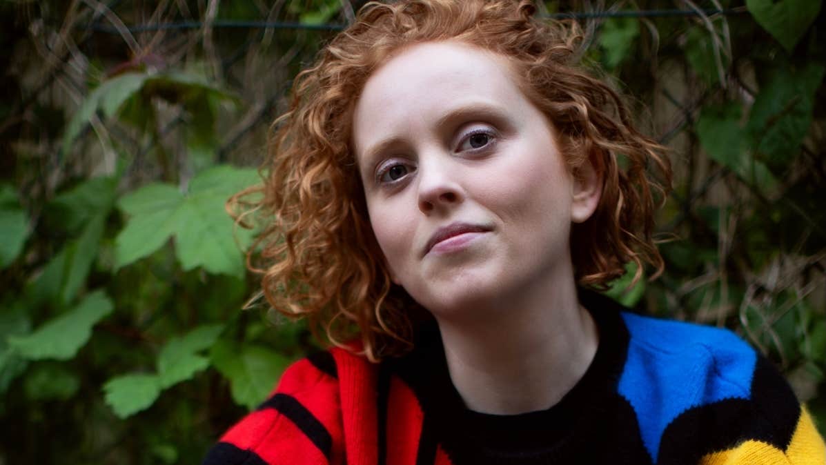 Jessie Grimes, presenter, gently smiling woman in colourful jumper against background of green plants