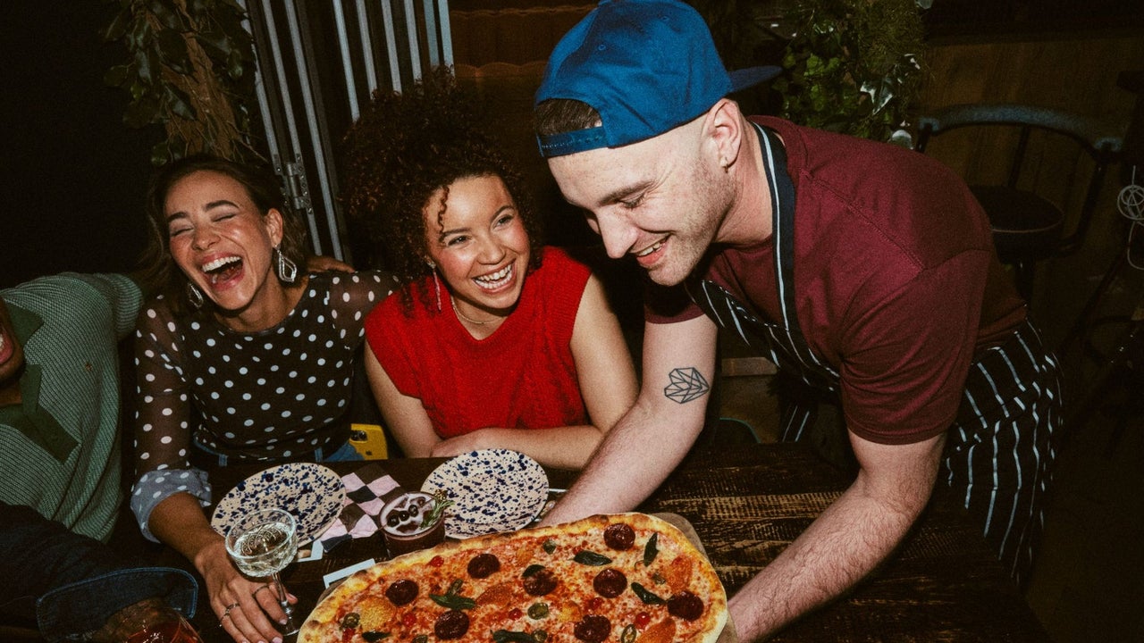 A waiter in an apron placing a large pizza on a table with smiling customers