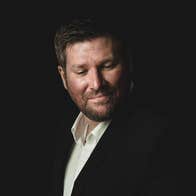 A man in dark jacket and white shirt is slightly smiling, looking down to his left, against dark background.