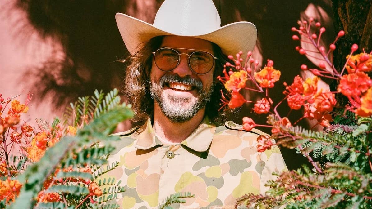 A wide smiling man in a cowboy hat, partially obscured by branches and foliage.