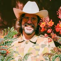 A wide smiling man in a cowboy hat, partially obscured by branches and foliage.