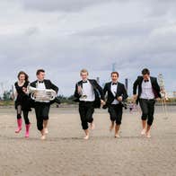 * musicians dressed running on Sandymount Strand with the Poolbeg towers and cloudy skyline behind them