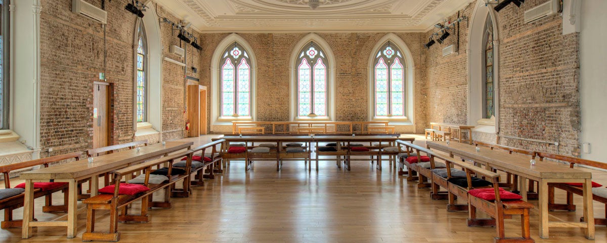 A large hall with rows of tables and seats along the wall with large Romanesque windows on the far wall