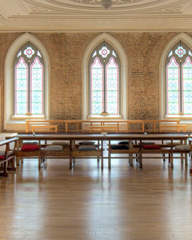 A large hall with rows of tables and seats along the wall with large Romanesque windows on the far wall