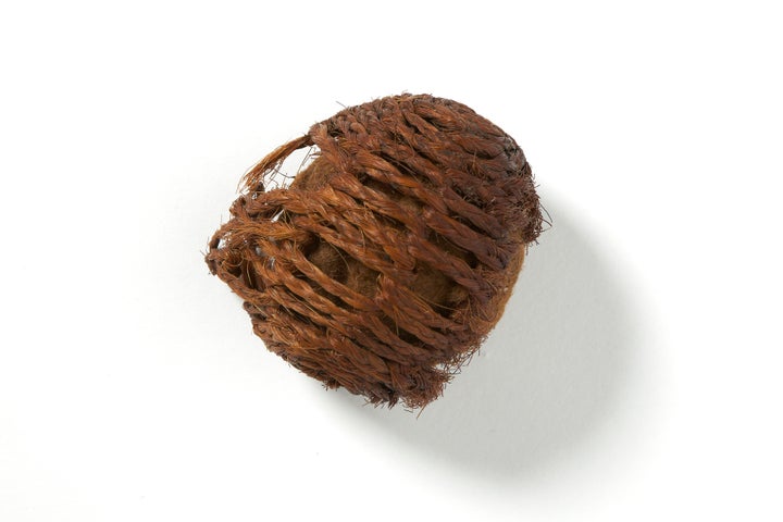 A small brown object in a rough ball shape made up of old interwoven twine, coming undone on one side, against a plain bright white background.