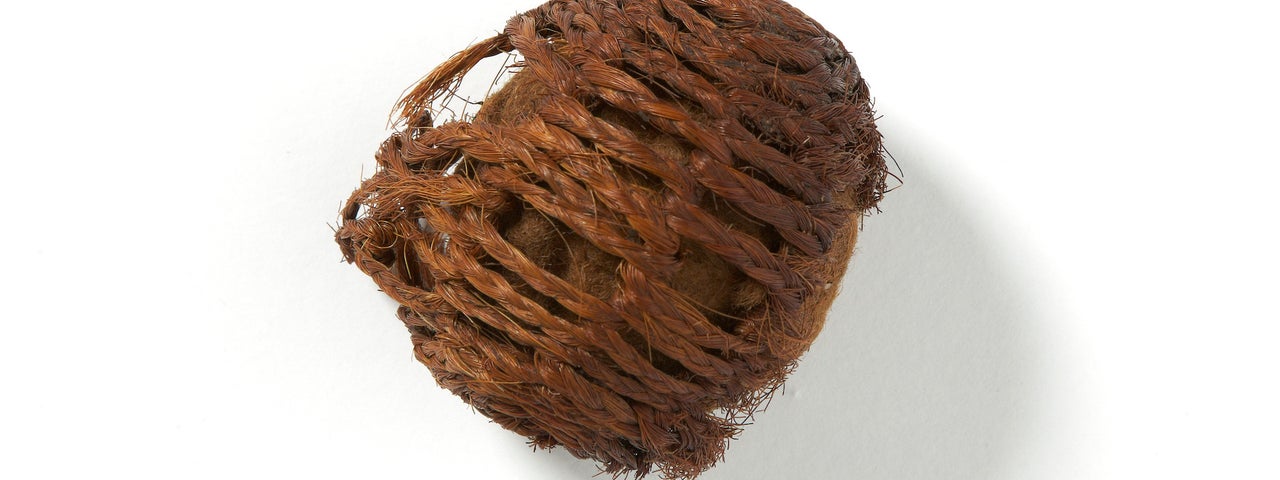 A small brown object in a rough ball shape made up of old interwoven twine, coming undone on one side, against a plain bright white background.