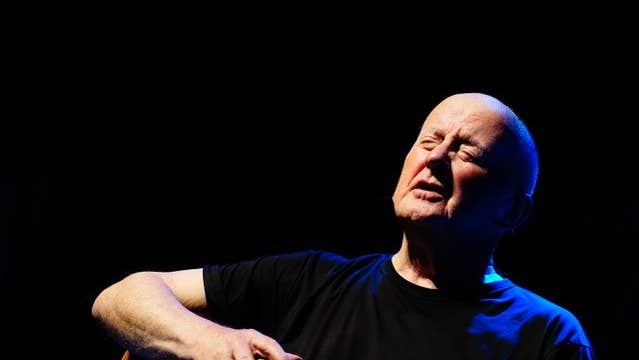 A bald man singing with eyes closed, holding a guitar.