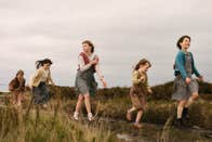 5 young girls run along the heather, on a boggy, mountainside path. Their clothes are old and woven, suggesting they are from the early 1900s in Ireland.