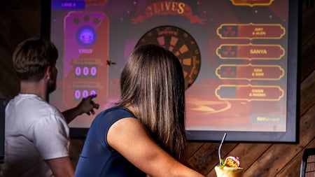 Person leaning on a counter with drinks as another person points at a digital dart board