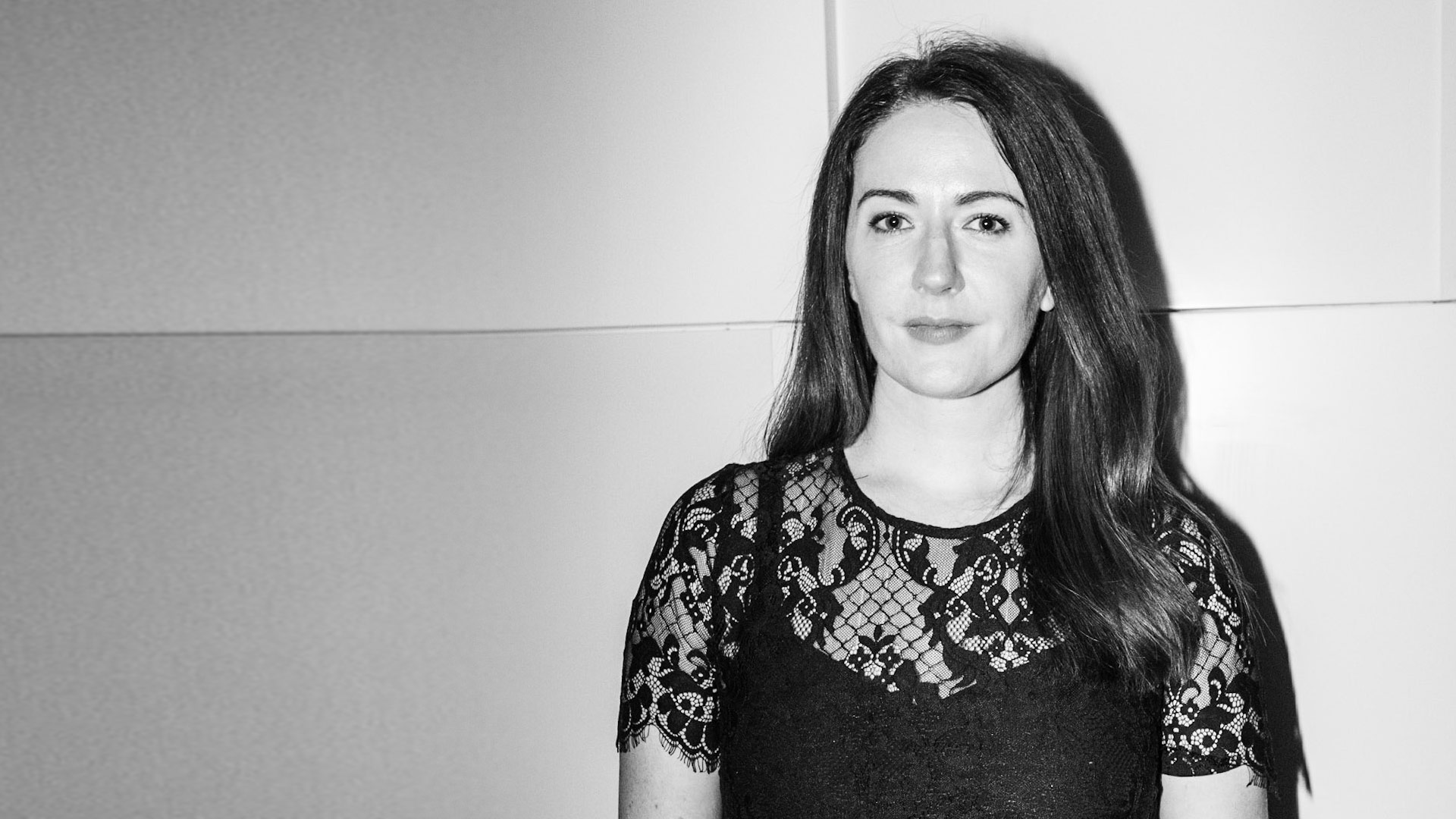 Black and white photo of woman in black lacy top, slight smile, against plain grey background.