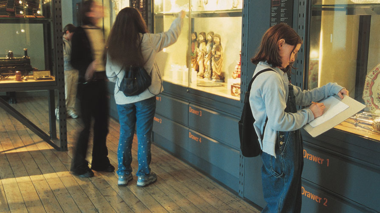 Children with paper and pens are in front of large display cabinets in a museum, other figures are blurred.
