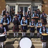 A group of people with drums dressed in blue kilts standing scattered around a flight of stone steps in front of a building