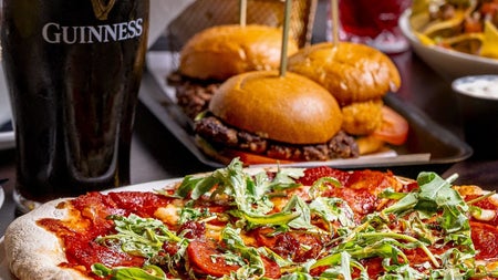 Pint of stout on a table next to a pizza sliders and a basket of chips in the background