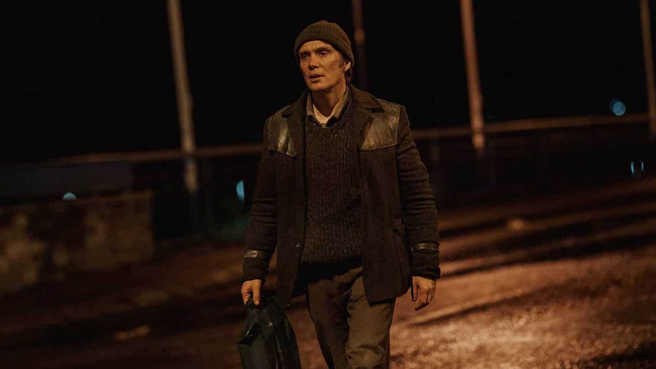 A man in well worn clothes is walking along a road at night.