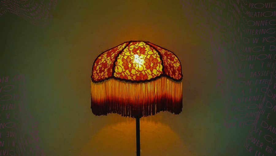 A foral lampshade with long, fringed edge, lit against dull green background.
