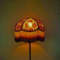 A foral lampshade with long, fringed edge, lit against dull green background.