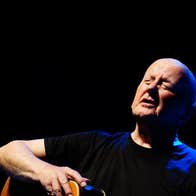 A bald man is singing with his eyes closed, a guitar on his lap.