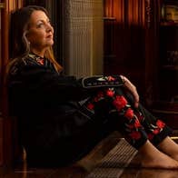A woman dressed in dark clothes with red flowers on is sat barefoot on a wooden floor, leaning against the wall, looking off into the distance.