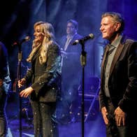 3 people standing with mics on a stage singing with band behind.