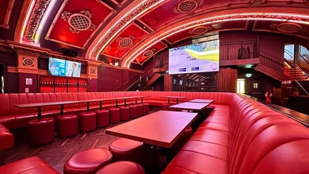 A theatre style lounge with luxurious red leather booth seating and ornate decorative ceilings with large screens throughout