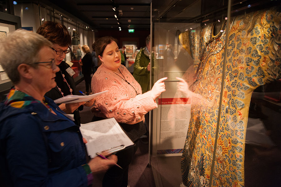A woman is talking and pointing at an ornate gown in a display cabinet, 2 women are standing close by listening and looking at clipboards and paper they are holding.