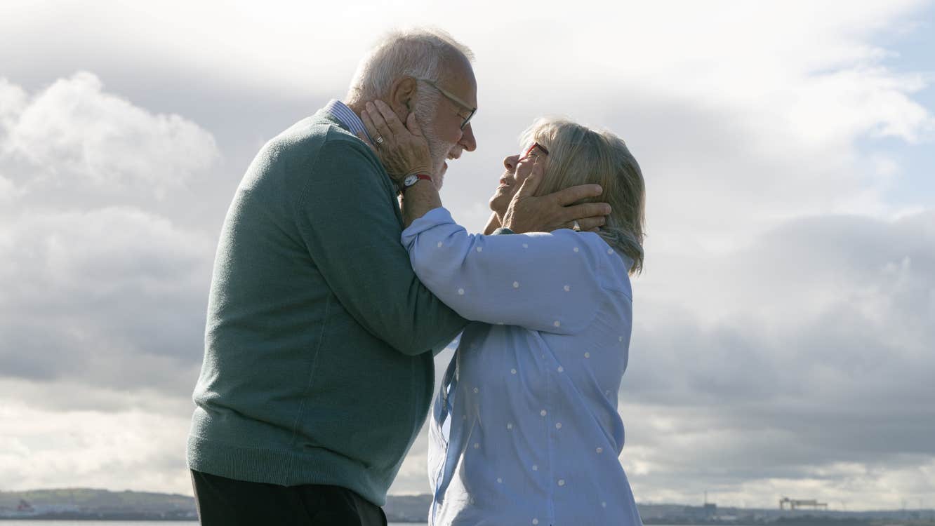 An old man and woman are facing each other, holding each other's heads tenderly.