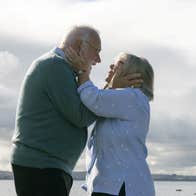 An old man and woman are facing each other, holding each other's heads tenderly.