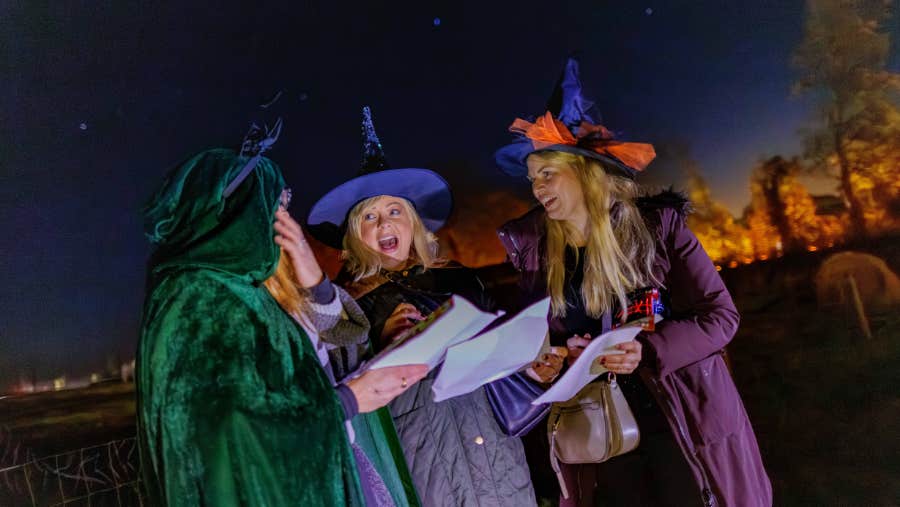 Adults in costume read their treasure hunt clues by torchlight at Nore Valley Park's Halloween Fire Trail, with warm lights glowing through the trees behind them.