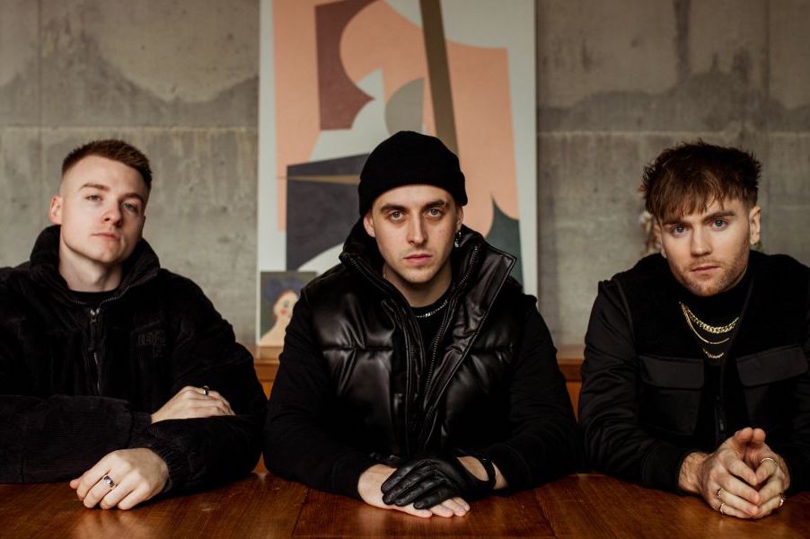 3 men dressed in black looking moody, sitting at a wooden table