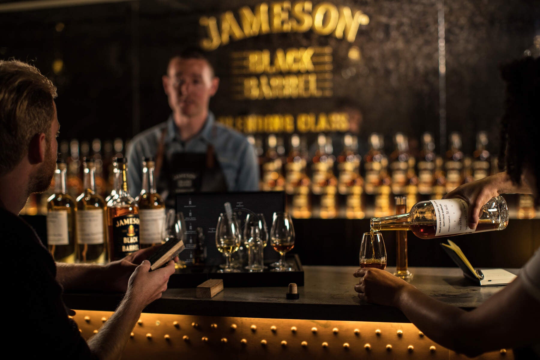Two people sitting at the bar in the Jameson Distillery while one person pours whiskey into a glass.