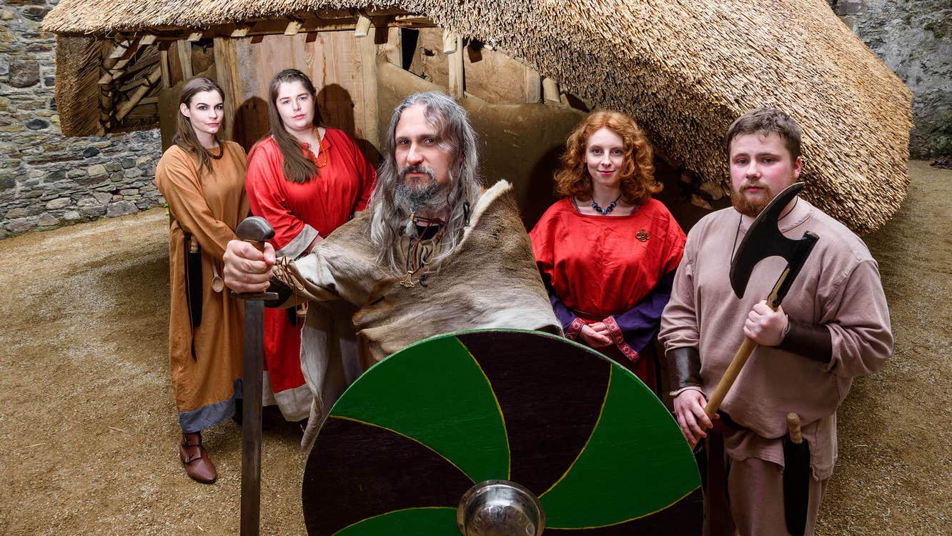 A group of Viking reenactors stand in front of a building carrying a sword, shield and axe.