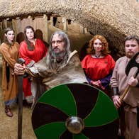 A group of Viking reenactors stand in front of a building carrying a sword, shield and axe.