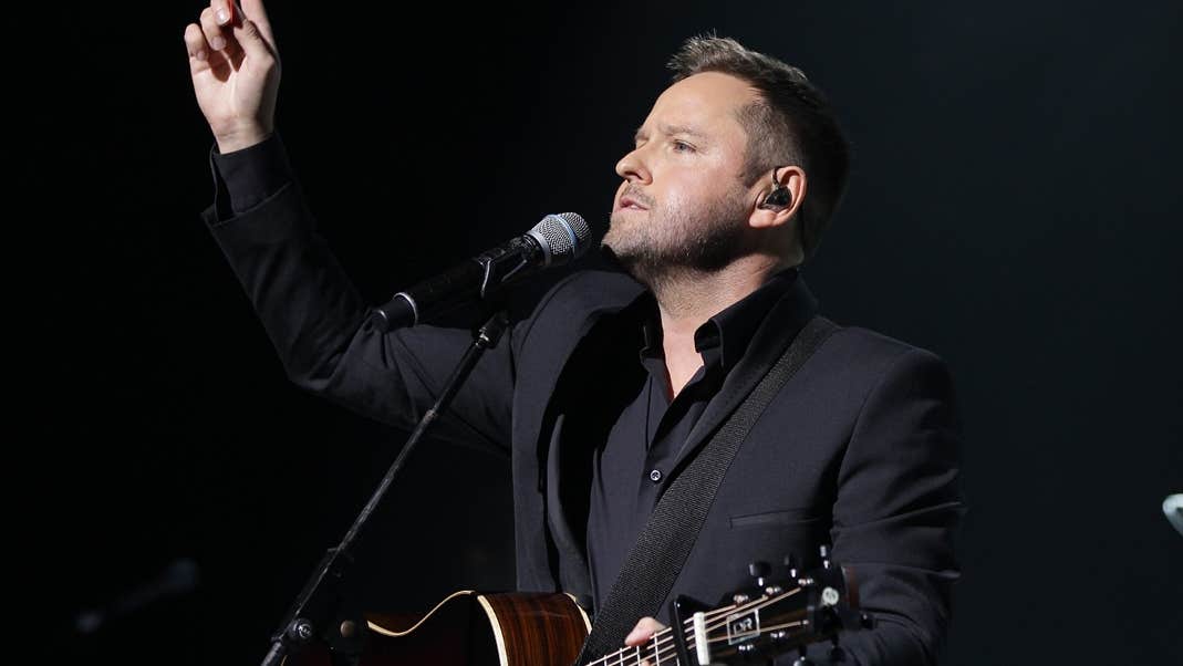 A man playing guitar with a mic on a stand and his right arm raised pointing up into the air.