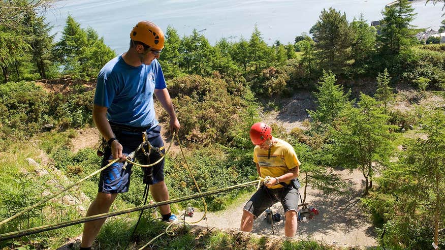 Man in blue using a rope to get down a slope with a person in a yellow tshirt