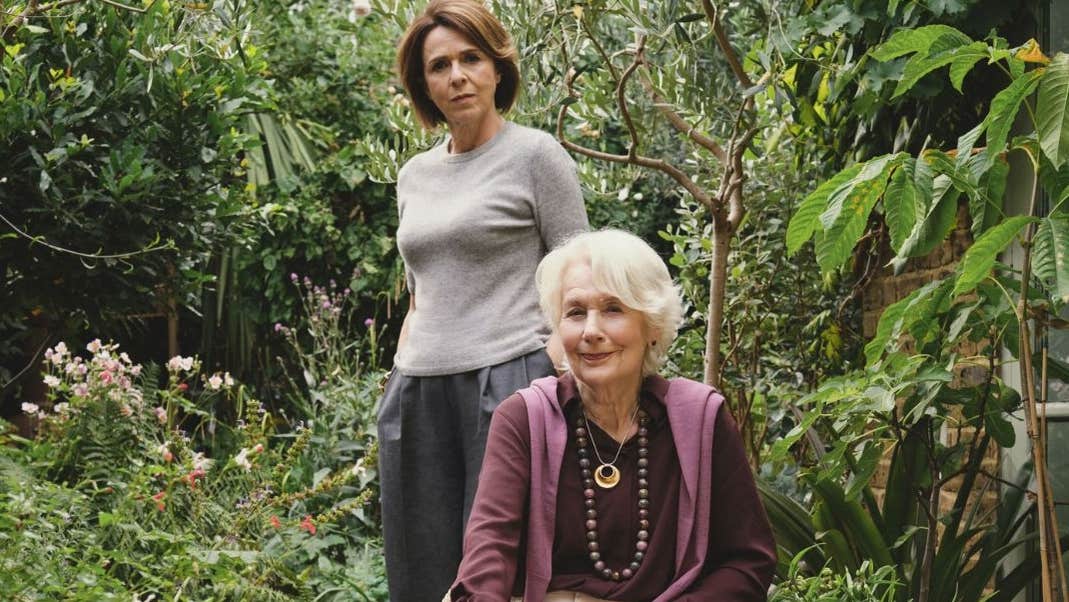 Meredith sits in a rattan chair in the garden, while her daughter Noelle stands behind her.